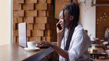 Young black woman talking on the phone while sitting in a cafe with a laptop video