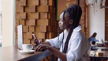giovane africano donna utilizzando Telefono mentre seduta nel un' bar con un' il computer portatile video