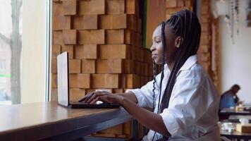 jovem africano americano mulher usando computador portátil computador dentro cafeteria. fêmea profissional trabalhando com caderno video