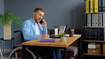 Motivated handicapped freelancer talking on the phone and taking notes sitting in wheelchair at office. Distant work and disability concept video