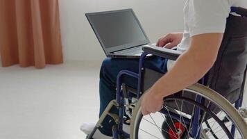 Disabled man sitting on wheelchair and using laptop in the living room. Freelancer, young writer or student work with laptop video