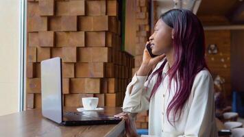 Young black woman talking on the phone while sitting in a cafe with a laptop video