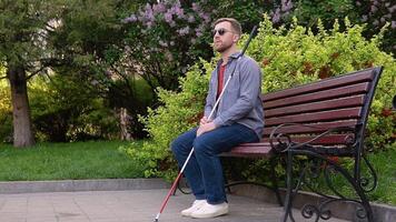 A blind man with a cane sits on a bench in a park video