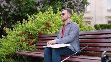 joven ciego hombre sentado en banco en ciudad parque y leyendo un braille libro video