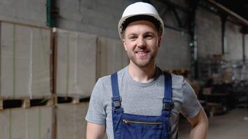 Handsome and happy professional worker in hat charmingly smiling on camera. In the background big warehouse video