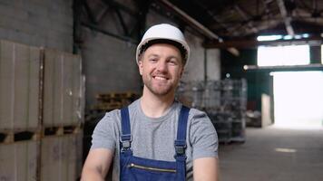 Handsome and happy professional worker in hat charmingly smiling on camera. In the background big warehouse video