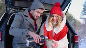 Young couple, a man and a woman, are sitting in the trunk of a car in a winter, snowy forest, hugging and drinking coffee video