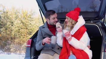 Young couple, a man and a woman, are sitting in the trunk of a car in a winter, snowy forest, hugging and drinking coffee video