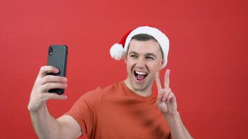 A man in Santa Claus hat expresses joy and good mood in the smartphone camera. A guy in red Christmas clothes with a smartphone in his hand is standing in front of a red background and make selfie video