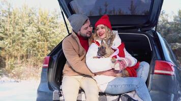 Young couple, a man and a woman with dog, are sitting in the trunk of a car in a winter and hugging, snowy forest video