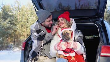 Young couple, a man and a woman with dog, are sitting in the trunk of a car in a winter and hugging, snowy forest video