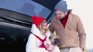 Young couple, a man and a woman with dog, are sitting in the trunk of a car in a winter and hugging, snowy forest video