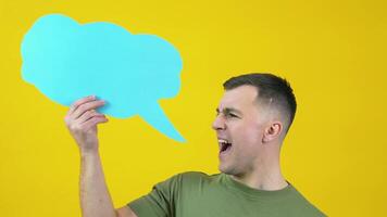 A man in a green T-shirt holds a cloud for insert text and shouts a message, slogan or song out loud. The guy is standing in front of a yellow background with a blue background for text in his hand video