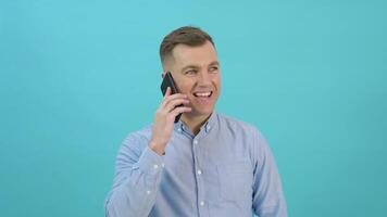 caucasien âge moyen homme dans une bleu chemise soulève une téléphone intelligent à le sien oreille et pourparlers. Bureau ouvrier ayant une téléphone conversation dans de face de une brillant bleu Contexte video