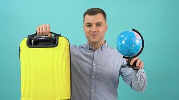 Happy positive man in a blue shirt raises a globe and a yellow suitcase to shoulder level and smiles broadly video