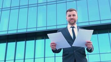 portret van een zelfverzekerd Mens in een bedrijf pak met documenten in voorkant van een modern kantoor gebouw video