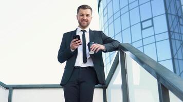 homem de negocios em pé lado de fora uma moderno corporativo construção com café copo e celular. paisagem urbana fundo video