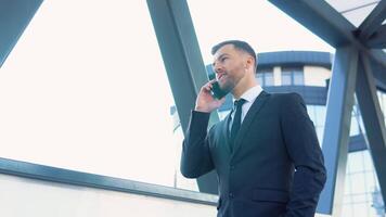 homem de negocios falando Móvel telefone perto moderno escritório prédio. retrato do feliz o negócio homem chamando telefone ao ar livre video