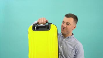 A caucasian man in a blue shirt expresses delight and joy with a yellow suitcase in his hands. An office worker is preparing for a vacation or business trip video