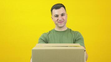 Young delivery man courier in a green T-shirt lifts a craft cardboard box in front of him. The guy is standing in front of a yellow background with a parcel in his hands video