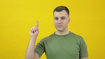 uma homem dentro uma verde camiseta levanta dele índice dedo acima para face nível. a cara é em pé dentro frente do uma amarelo fundo video
