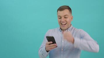 une homme dans une bleu chemise est correspondant sur une téléphone intelligent et exprime génial joie et surprendre. Bureau ouvrier avec une mobile téléphone dans le sien mains des stands dans de face de une brillant bleu Contexte video