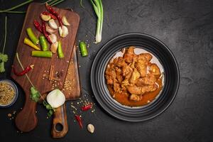 Delicious beef or pork stew in a rustic black plate with fresh coriander, red chilies, garlic, onions and spices placed on a black background, top view photo