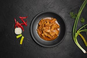 Delicious beef or pork stew in a rustic black plate with fresh coriander, red chilies, garlic, onions and spices placed on a black background, top view photo