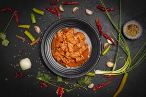 Delicious beef or pork stew in a rustic black plate with fresh coriander, red chilies, garlic, onions and spices placed on a black background, top view photo