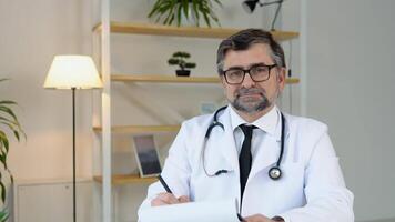 Sénior médecin dans blanc costume avec stéthoscope dans le sien médical bureau. vieux mature médecin thérapeute travail à Bureau video