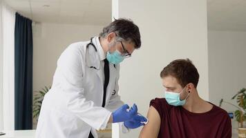 Senior doctor giving Covid-19 or flu antivirus vaccine shot to young male patient wear face mask protection from virus disease at health clinic or hospital office. Vaccination concept video