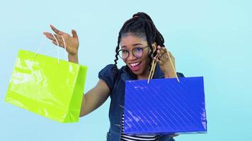 africano americano ragazza nel blu Abiti Tenere laminato carta borse con penne. adolescente ragazza in piedi su un' solido leggero blu sfondo video