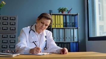 Senior female doctor writing information in medical registration journal. Old mature doctor therapist working at office video