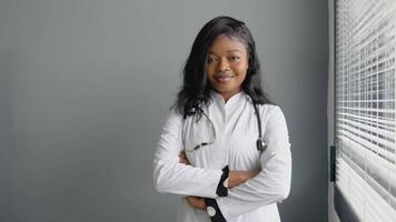 retrato de hermoso africano hembra médico vistiendo blanco Saco y estetoscopio cruces brazos, en pie en su salud clínica oficina video