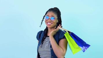 Cute cheerful young african american girl in blue clothes holding laminated paper bags with pens. Teen girl standing on a solid light blue background video