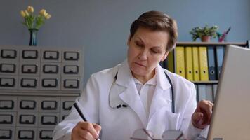 Senior woman doctor 50s works with a laptop and writes down the patient's medical history video