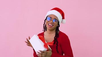 Young african american girl has fun with a gift. Female student in red clothes and Santa's hat holds a white box with a red ribbon video
