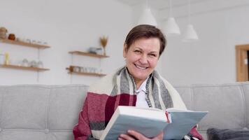 Senior woman reading a book while sitting on the couch at home. Retirement lifestyle of woman self isolating in quarantine lockdown video