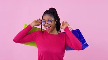 Cute cheerful young african american girl in pink clothes having fun and dancing with paper laminated bags with handles. Teen girl standing on a solid pink background video