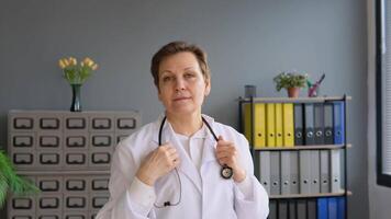 positiv allmän praktiker stänga upp ansikte porträtt. Lycklig senior europeisk kvinna läkare bär vit medicinsk täcka och stetoskop ser på kamera video