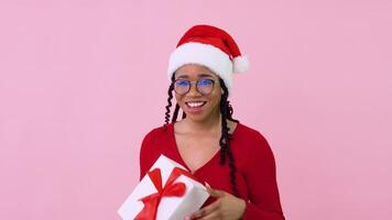 fofa jovem africano americano menina mostra grande dedo acima com uma presente. fêmea aluna dentro vermelho roupas e papai noel chapéu detém uma branco caixa com uma vermelho fita video