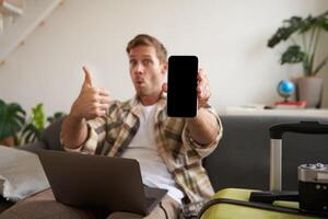 Portrait of handsome young man, showing smartphone screen, recommending mobile app for travelling or booking online, sitting with laptop and suitcase photo