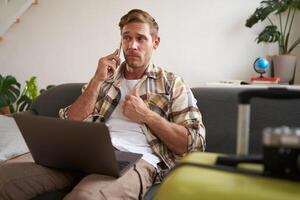 Portrait of man talking on mobile phone, tourist with laptop and suitcase answers telephone photo