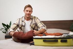 Portrait of handsome young man, tourist unpacking clothes in hotel room, staying in hostel on vacation, travelling, sitting with luggage on bed photo