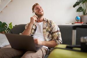 Portrait of young smiling man with suitcase and laptop, making a phone call, booking a flight tickets, calling to confirm hotel room photo