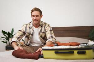 Portrait of young man, traveller unpacking suitcase, putting clothes on bed, smiling, enjoying vacation, hotel stay photo