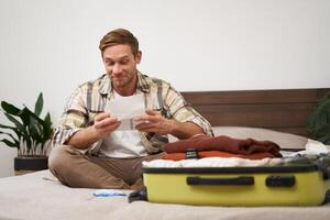 Portrait of excited young man, tourist going on vacation, has his tickets, sitting near opened suitcase, looking at passport with pleased smile photo