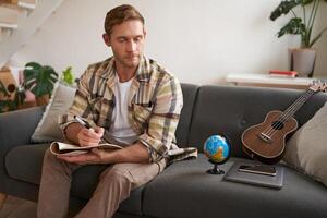 Man writing check-list, packing for holiday, cross-reference items with his notes, sits in living room with globe and ukulele photo