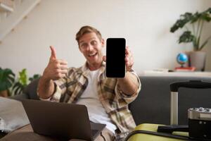 Portrait of man, tourist shows his mobile phone screen, recommends travel app, sitting with suitcase and laptop photo