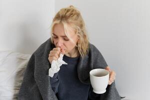 Concept of flu and quarantine. Close up portrait of young woman feeling sick, caught a cold, sneezing and blowing nose in napkin, holding warm tea in hand photo